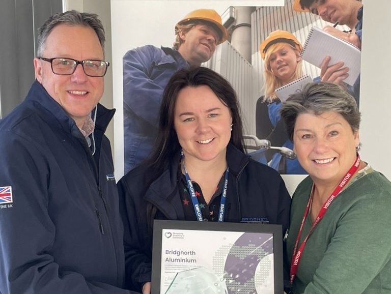 Three people pose together holding a framed certificate or award relating to Bridgnorth Aluminium. A man in a dark jacket and glasses stands on the left, a woman in a dark jacket with a lanyard stands in the centre, and a woman in a green jumper with a red lanyard stands on the right. Behind them is a large display banner showing the Bridgnorth Aluminium company branding and images of workers in orange hard hats at what appears to be an industrial facility.