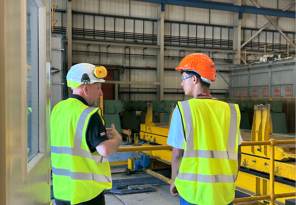 Experienced factory worker in hard hard and hi-viz jacket talks to young worker, possibly on an apprenticeship, also in PPE