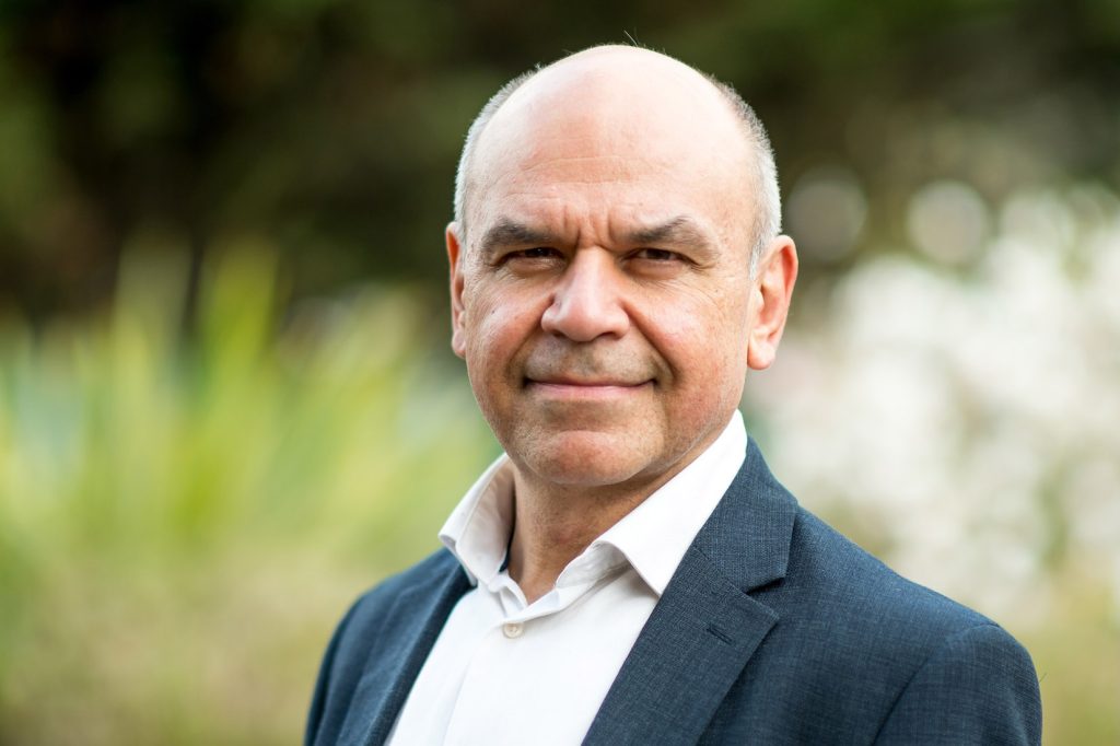 A person wearing a suit, no tie, shirt unbuttoned stood against a blurred out natural green background showing plants.