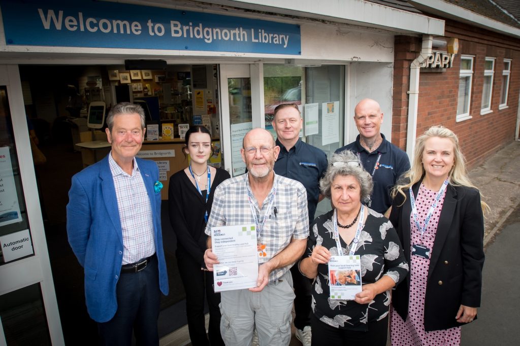 A group of people stood outside a community library. Two of the are holding up pieces of paper.