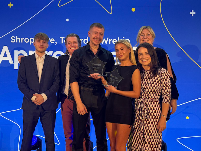 Six people stood together at an awards ceremony. One of them is holding two awards.