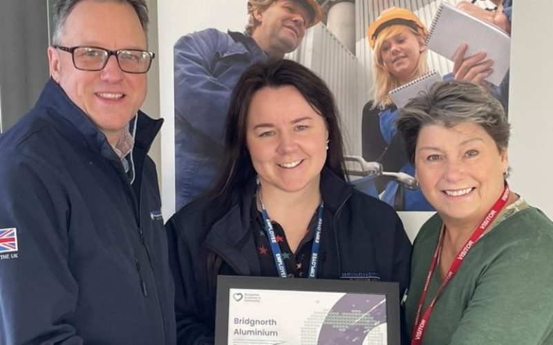 Three people pose together holding a framed certificate or award relating to Bridgnorth Aluminium. A man in a dark jacket and glasses stands on the left, a woman in a dark jacket with a lanyard stands in the centre, and a woman in a green jumper with a red lanyard stands on the right. Behind them is a large display banner showing the Bridgnorth Aluminium company branding and images of workers in orange hard hats at what appears to be an industrial facility.