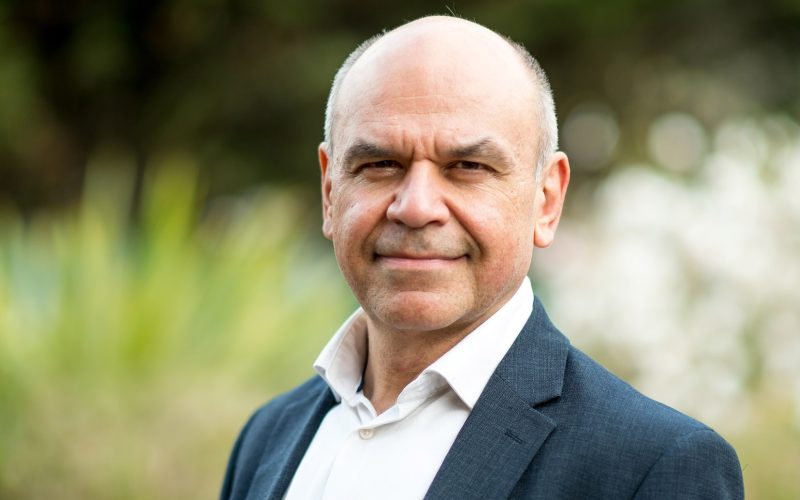 A person wearing a suit, no tie, shirt unbuttoned stood against a blurred out natural green background showing plants.