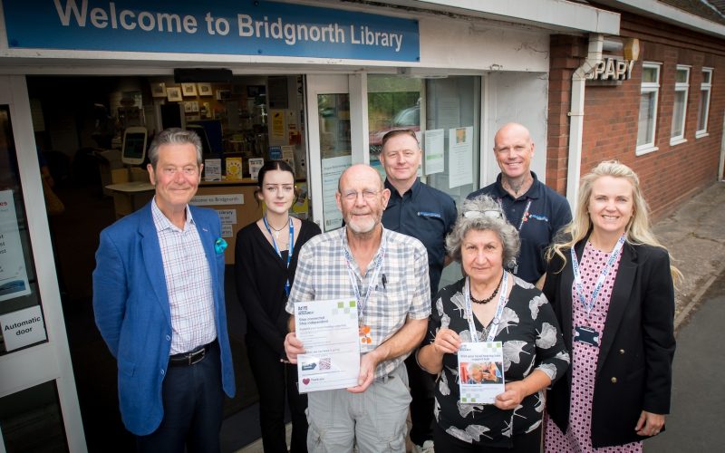 A group of people stood outside a community library. Two of the are holding up pieces of paper.