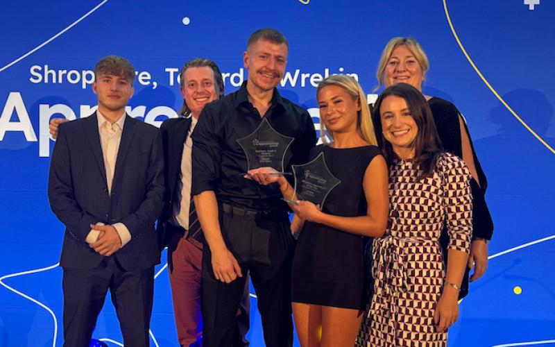 Six people stood together at an awards ceremony. One of them is holding two awards.